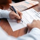 Person reviewing and writing on financial documents with a pen on a wooden desk.